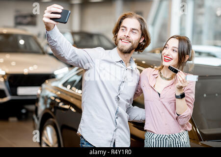 Coppia felice rendendo selfie foto tenendo premuto i tasti nella parte anteriore della loro nuova automobile nello showroom Foto Stock