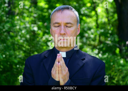 Guardare un buon sacerdote cattolico è pregare nella foresta. Egli si unisce a mani. Gli occhi chiusi. Foto Stock