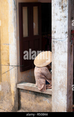 Donna anziana nel tradizionale abbigliamento vietnamiti seduti sul muro di pietra che indossa la paglia coolie hat in Hoi An Old Town, Quang Nam Provincia, Vietnam Foto Stock