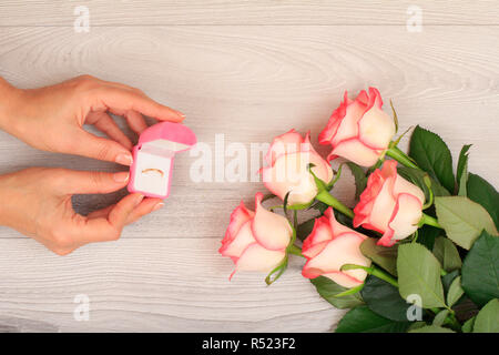 Donna di mani tenendo una scatola con anello d'oro su legno grigio Sfondo con bellissime rose. Concetto di dare un regalo per le vacanze. Vista dall'alto. Foto Stock