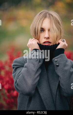 Foto di giovane donna in cappotto grigio su sfondo sfocato di piante rosse della città Foto Stock