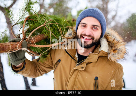 Uomo Barbuto che trasportano appena tagliato all albero di natale nella foresta. I giovani orsi lumberjack abete sulla sua spalla nei boschi. Un comportamento irresponsabile verso la natura, salvare la foresta, mantenere il concetto di verde. Foto Stock