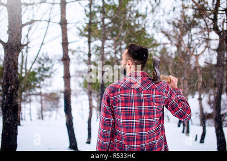 Tanga barbuto uomo in un inverno di boschi innevati con ax su una spalla. Woodman in piedi nella foresta. Maschio di ispezione degli alberi nei boschi. Lumberjack taglialegna holding ax indossando plaid camicia rossa. Foto Stock