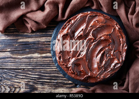 In casa la torta al cioccolato a strati con confettura di albicocca e guarnita con glassa al cioccolato su una pietra nera la piastra con un panno marrone su un vecchio rustico in legno tab Foto Stock