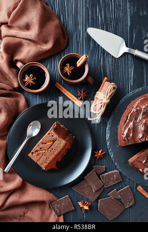 Una fetta di torta al cioccolato con marmellata di albicocche il riempimento e rabboccato con glassa al cioccolato servito su una piastra con vin brulé in argilla tazze rustico su un nero woo Foto Stock