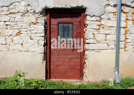 Nuove porte in legno formato da pannelli con ferro forgiato latch e finestra di barre di protezione montato su fatiscenti incrinato muro di pietra accanto alla nuova acqua grondaia Foto Stock