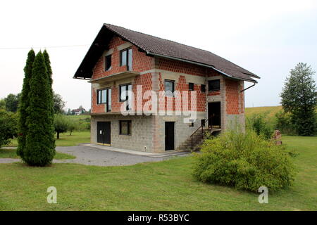 Incompiuta mattone rosso e grigio building block famiglia casa suburbana con nuove porte e finestre circondate con erba appena tagliata e piccoli alberi sulla guerra Foto Stock
