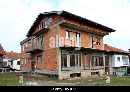 Incompiuta di mattoni rossi suburban casa familiare con imbarcati finestre e porte anteriori circondato da erba e altre case su freddo giorno di primavera Foto Stock