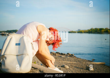 La ragazza di colore rosa pallido vestito con i capelli rossi e uno zaino camminando lungo la riva del fiume, seduto sulla riva sabbiosa Foto Stock