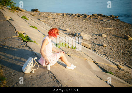 La ragazza di colore rosa pallido vestito con i capelli rossi e uno zaino camminando lungo la riva del fiume, seduto sulla riva sabbiosa Foto Stock
