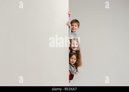 Banner con sorpresa di vedere i bambini sbirciare il bordo con copyspace. Il ritratto del piccolo grazioso ragazzo di ragazzi e ragazze guardando la telecamera contro studio bianco parete. Moda per bambini e felice concetto emozioni Foto Stock