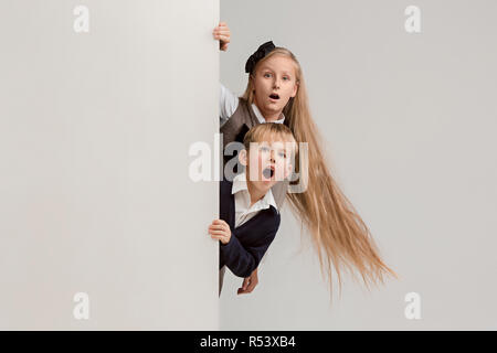 Banner con sorpresa di vedere i bambini sbirciare il bordo con copyspace. Il ritratto del piccolo grazioso ragazzo di ragazzi e ragazze guardando la telecamera contro studio bianco parete. Moda per bambini e felice concetto emozioni Foto Stock