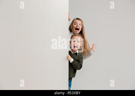 Banner con sorpresa di vedere i bambini sbirciare il bordo con copyspace. Il ritratto del piccolo grazioso ragazzo di ragazzi e ragazze guardando la telecamera contro studio bianco parete. Moda per bambini e felice concetto emozioni Foto Stock