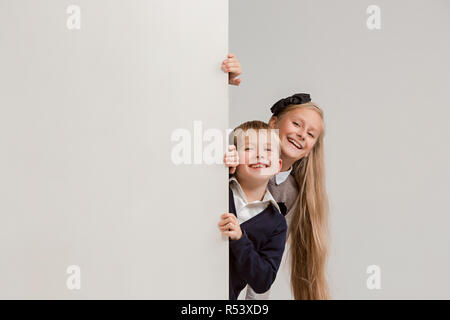 Banner con sorpresa di vedere i bambini sbirciare il bordo con copyspace. Il ritratto del piccolo grazioso ragazzo di ragazzi e ragazze guardando la telecamera contro studio bianco parete. Moda per bambini e felice concetto emozioni Foto Stock