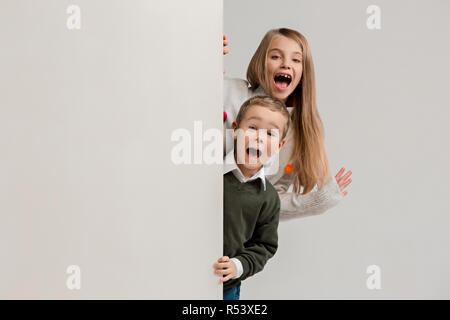 Banner con sorpresa di vedere i bambini sbirciare il bordo con copyspace. Il ritratto del piccolo grazioso ragazzo di ragazzi e ragazze guardando la telecamera contro studio bianco parete. Moda per bambini e felice concetto emozioni Foto Stock