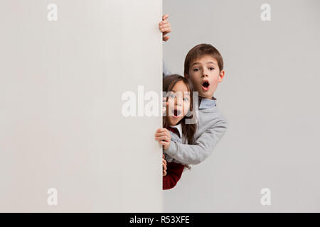 Banner con sorpresa di vedere i bambini sbirciare il bordo con copyspace. Il ritratto del piccolo grazioso ragazzo di ragazzi e ragazze guardando la telecamera contro studio bianco parete. Moda per bambini e felice concetto emozioni Foto Stock