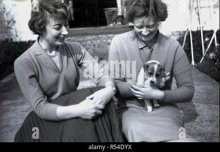 Degli anni Cinquanta, due eleganti e ben dresse giovani donne, uno in gonna plissettata e superiore di lana - possibilmente sorelle - seduti insieme in un giardino sul retro, uno tenendo un piccolo cane, un jack russell terrier, Inghilterra, Regno Unito. Foto Stock