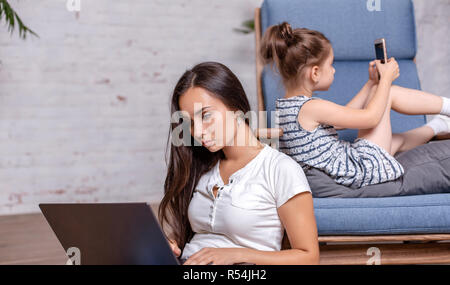 La madre lavora con laptop, seduto sul pavimento e graziosa bambina posa sulla poltrona e la riproduzione di videogiochi sullo smartphone Foto Stock