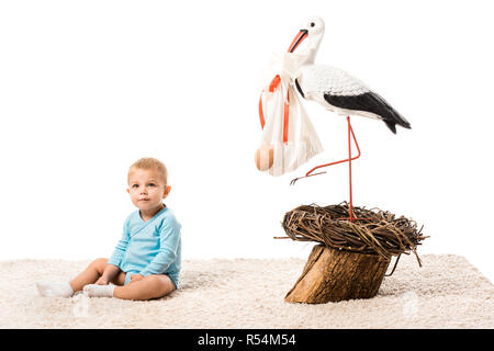 Adorabili toddler boy in blue body seduta sul tappeto vicino al grande cicogna decorativo isolato su bianco Foto Stock