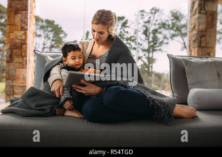 Madre e figlio seduti sul divano avvolto in una coperta guardando digitale compressa. Ragazzino con la madre guardare cartoni sul tablet pc in living roo Foto Stock