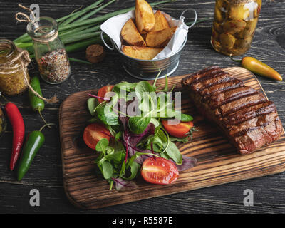 Bistecca su scheda con erbe aromatiche, patate fritte, spezie su una superficie nera Foto Stock
