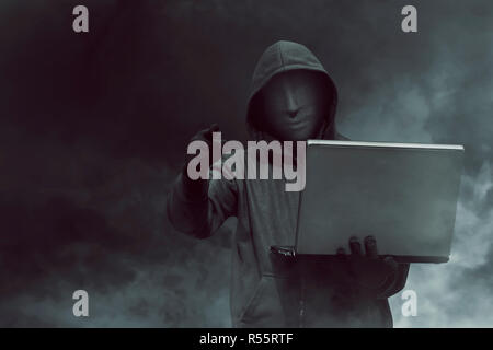 Ritratto di hacker con cappuccio con maschera holding laptop mentre in piedi Foto Stock