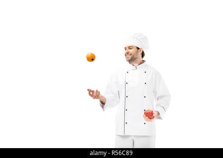 Sorridente giovane chef giocoleria con pomodoro e peperone isolato su bianco Foto Stock