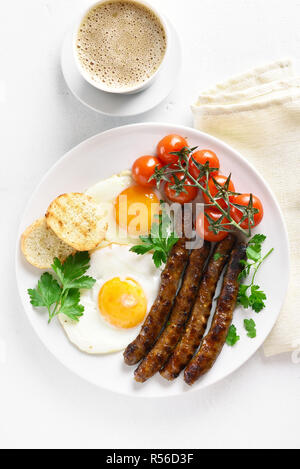Uova fritte, salsicce, pomodori ciliegia sulla piastra e la tazza di caffè sulla pietra bianca dello sfondo. Vista superiore, flay lay Foto Stock