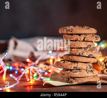 Round biscotti al cioccolato in una pila, intorno al nuovo anno di ghirlande di masterizzazione su un tavolo marrone Foto Stock