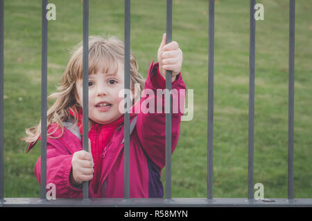 Poco carino Caucasian ragazza vestita in camicia rosa in piedi dietro i cancelli di metallo che conduce al parco giochi Foto Stock