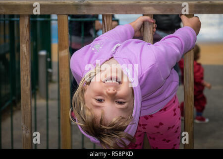 Poco carino Caucasian ragazza vestita in rosa hoodie mantenendo la recinzione di legno barre sul parco giochi Foto Stock