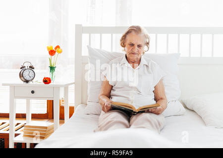 Donna anziana seduti comodamente nel letto leggendo il suo libro preferito Foto Stock