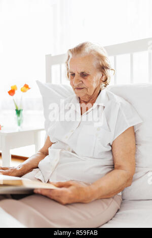 Donna anziana seduti comodamente nel letto leggendo il suo libro preferito Foto Stock