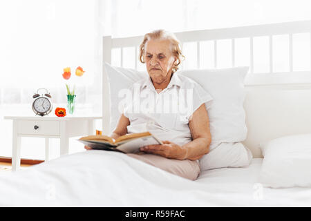 Donna anziana seduti comodamente nel letto leggendo il suo libro preferito Foto Stock