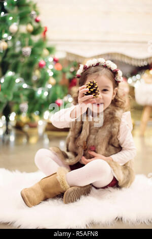 Carino bambina festeggia il Natale a casa. Si siede su una sedia e detiene un cono di abete nelle sue mani. Emozioni positive. Albero di natale in backgro Foto Stock