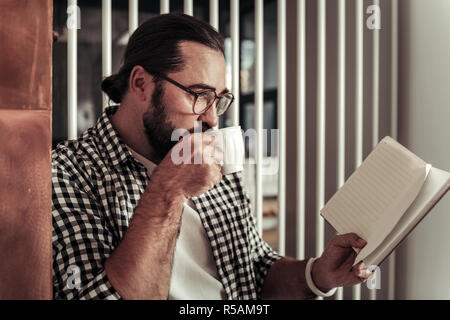 Drink di mattina. Nizza uomo positivo prendendo un sorso di caffè durante la lettura del suo notebook Foto Stock