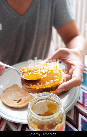 Primo piano di un giovane uomo caucasico, indossando un grigio casual T-shirt, seduti a un tavolo set, diffondendo alcuni arancione o confettura di pesche su un toast Foto Stock