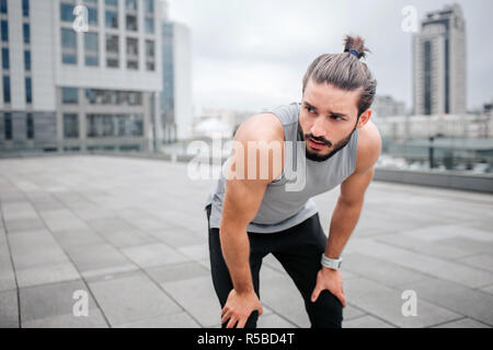 Stanco giovane uomo ha il resto. Lui si alza e si appoggia sulle gambe con le mani. Guy guarda a destra con grave vista. Egli è la calma. Giovane uomo si erge solitaria su strada Foto Stock