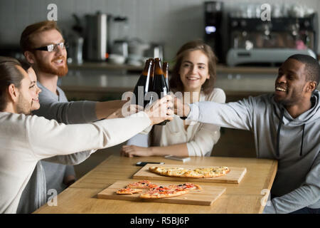 Diversi amici divertirsi in cafe toast dire ciao, multietnica agli studenti di mangiare pizza e bere birra, il tintinnio delle bottiglie eccitato celebrando riunioni, millen Foto Stock