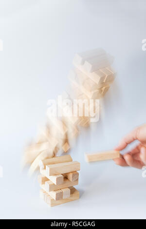 Mano d'uomo prendendo un blocco da una torre instabile di blocchi di legno facendolo cadere. Concetto foto di assunzione di rischi e la mancanza. Sfondo grigio chiaro Foto Stock