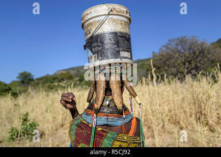 Immagine da dietro di una donna Muila con ornamenti tradizionali e taglio di capelli che porta un secchio pieno di acqua. Foto Stock