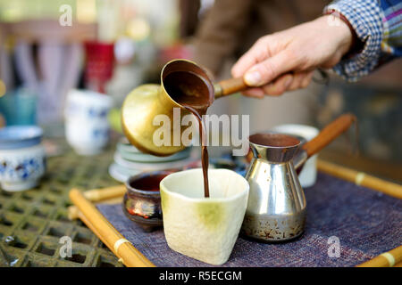 Versare il caffè caldo da un rame cezve nella tazza di ceramica in accogliente cucina. In ceramica colorata e tazze di caffè caldo. Preparare un delizioso caffè Foto Stock