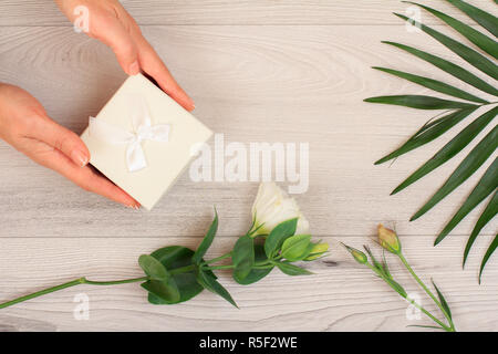 Donna di mani tenendo una confezione regalo su grigio Sfondo di legno con splendidi fiori e verde leavs. Concetto di dare un regalo in vacanza o compleanno. Parte superiore Foto Stock