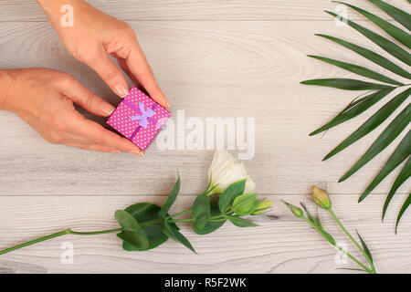 Donna di mani tenendo una confezione regalo su grigio Sfondo di legno con splendidi fiori e verde leavs. Concetto di dare un regalo in vacanza o compleanno. Parte superiore Foto Stock