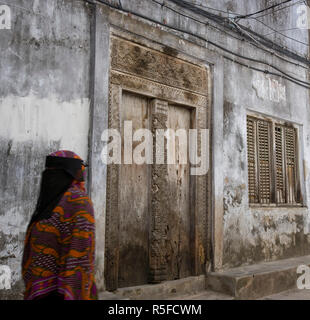 Zanzibar, Tanzania. Un Zanzibari donne nei tradizionali Khanga accappatoi, passando una vecchia porta di legno in Stone Town. Foto Stock