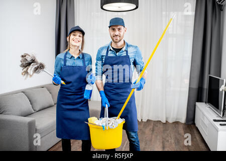 Ritratto di una giovane coppia come pulitori professionali in uniforme blu con gli strumenti di pulizia in appartamento Foto Stock