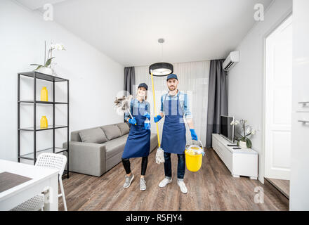 Ritratto di una giovane coppia come pulitori professionali in uniforme blu con gli strumenti di pulizia nell'appartamento. Ampio angolo di visione Foto Stock