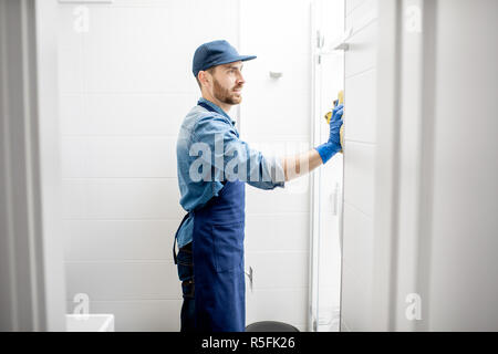 L uomo come pulitore professionale tergi la porta della doccia con tergicristallo di cotone nel bagno bianco Foto Stock