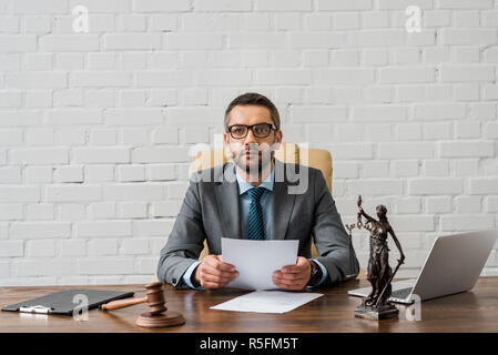 Giudice serio in occhiali a lavorare con documenti e guardando la telecamera in ufficio Foto Stock