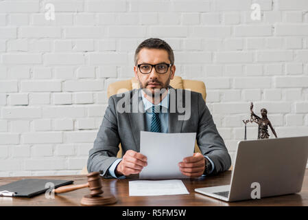 Giurista serio in occhiali a lavorare con documenti e guardando la telecamera in ufficio Foto Stock